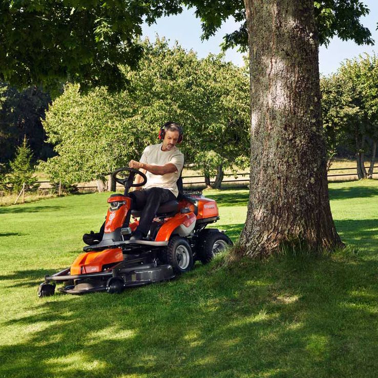 Ein Mann fährt konzentriert einen roten Aufsitzrasenmäher auf einer grünen Wiese unter Bäumen.