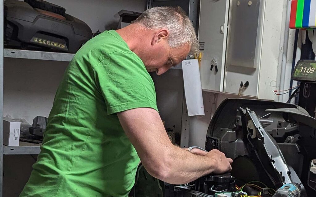 Älterer Mann in grünem Shirt repariert konzentriert ein Gerät in Werkstatt. Beige Wand im Hintergrund.